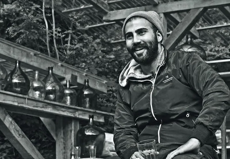 Ömer Kaya mit Mütze und Jacke lachend an einem Holztisch, schwarzweiß