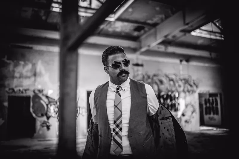 Ömer Kaya mit Weste und Sonnenbrille in einer verfallenen Halle, schwarzweiß