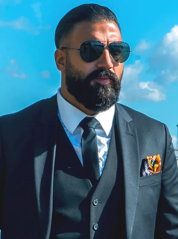 Ömer Kaya im grauen Dreiteiler mit Einstecktuch und Pilotenbrille vor blauem Himmel