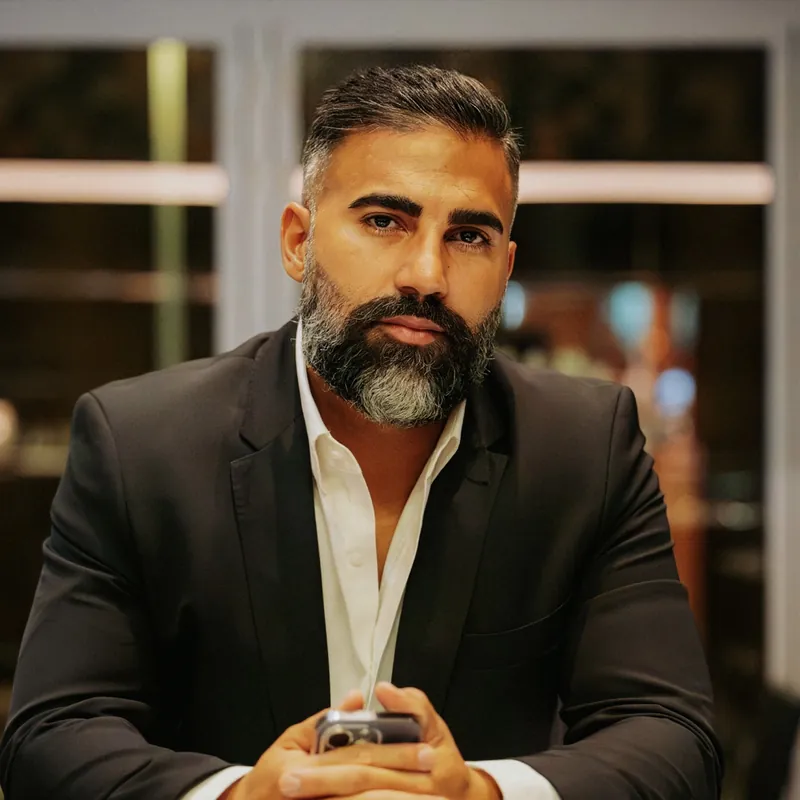 Ömer Kaya im schwarzen Sakko mit grauem Bart, das Telefon in den gefalteten Händen